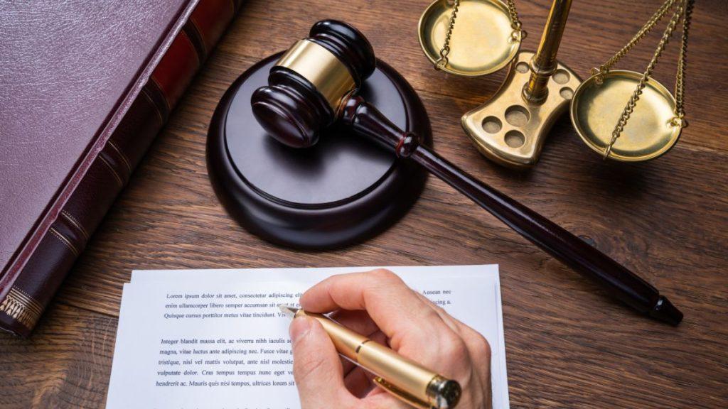 Close-up of a legal desk: a gavel, balance scales, a binder, and a hand signing a document on wood grain.