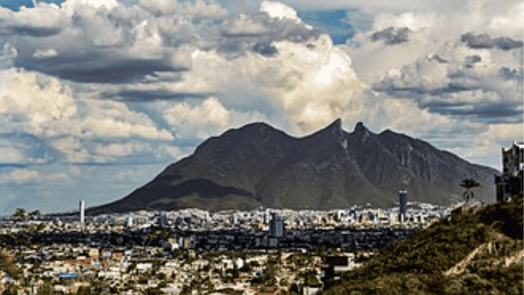 City skyline at the base of a large, jagged mountain under a cloudy sky.