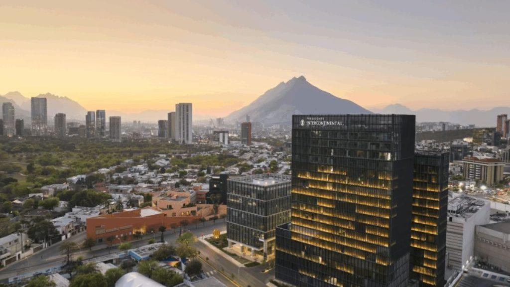 A city skyline at dusk with a tall glass InterContinental hotel in the foreground and a mountain range in the background, softly lit by sunset light.