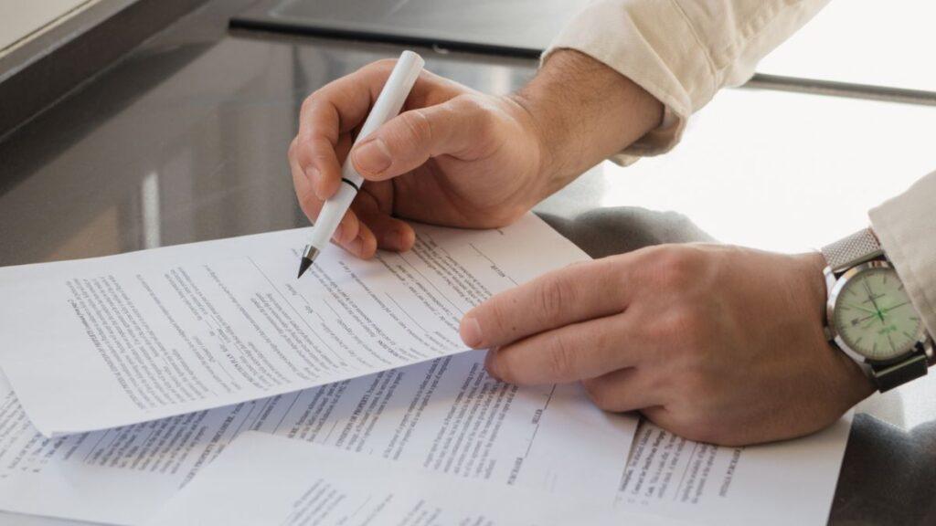 bróker-inmobiliario-quantium-desarrollos-contrato - CLAVE NOTICIA Person signs a contract with a white pen over stacked documents on a glass desk, wearing a silver wristwatch.