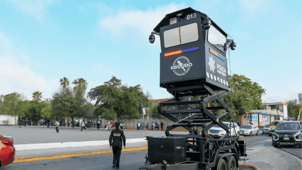 Mobile police surveillance platform mounted on a scissor-lift trailer on a sunny city street, officer nearby.