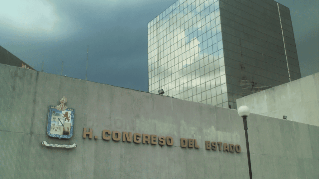 Exterior view of a government building with the inscription H. Congreso del Estado and a coat of arms, against a cloudy sky and a glass high-rise in the background.