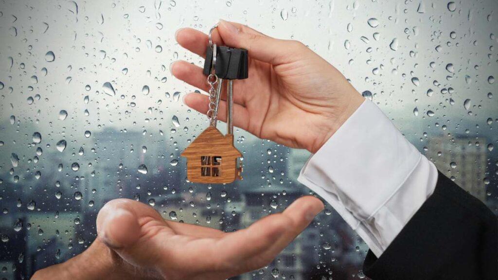 Imágen de unas manos sosteniendo un llavero con una figura de una casa pequeña y al fondo una ventana que refleja el exterior con un clima lluvioso
