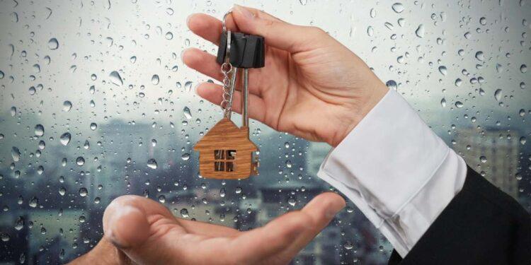 Imágen de unas manos sosteniendo un llavero con una figura de una casa pequeña y al fondo una ventana que refleja el exterior con un clima lluvioso